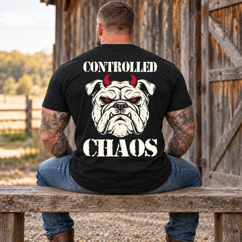 Man wearing a black CONTROLLED CHAOS veteran T-shirt, back print view, wooden bench by a weathered barn