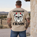 Man wearing a sand CONTROLLED CHAOS veteran T-shirt, back print view, stone wall by a ranch fence