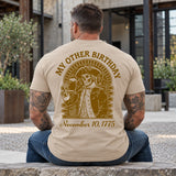 Man wearing a sand My Other Birthday custom year veteran T-shirt, back print view, stone courtyard outside a modern building