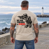 Man wearing a sand We Run Towards the Gunfire custom year veteran T-shirt, back print view, stone pier facing a lighthouse